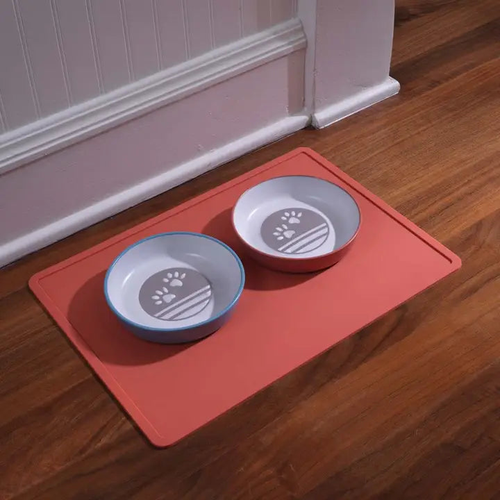 Two pet bowls with paw prints on a red mat on a wooden floor.