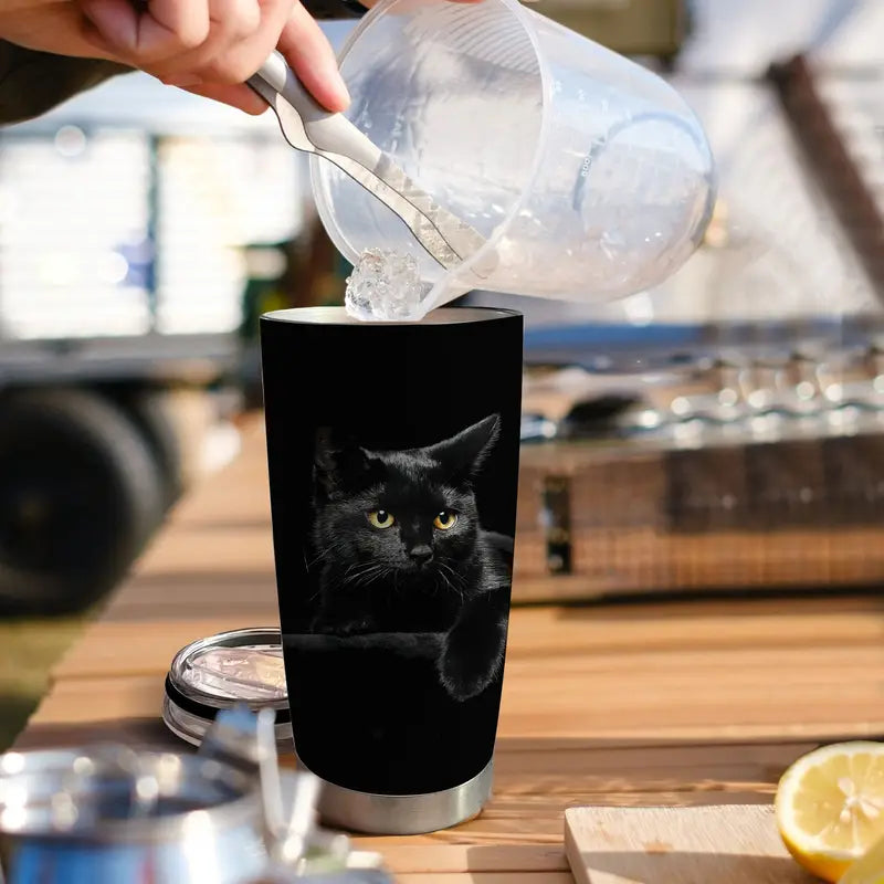 Person pouring liquid into a tumbler with a black cat design on a wooden table.