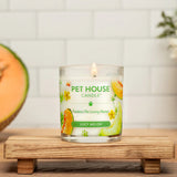 Pet House Candle labeled 'Juicy Melon' on a wooden surface with a cantaloupe and flowers in the background.