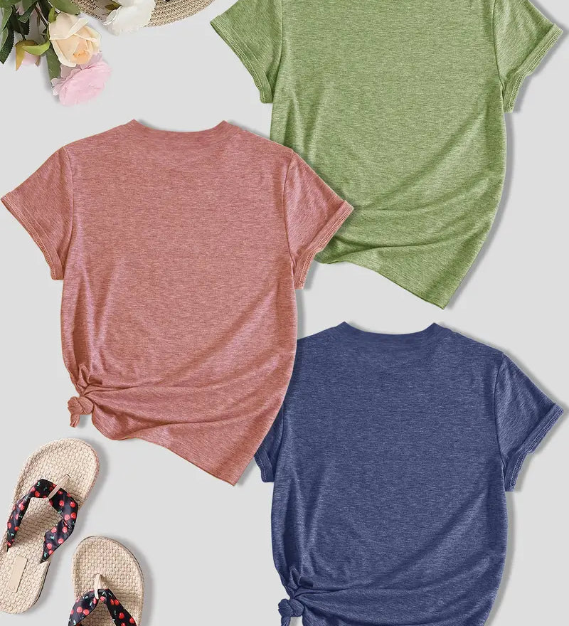 Three t-shirts in pink, green, and blue on a light surface with flip-flops and flowers.