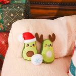 Two avocado-shaped toys with Christmas hats and antlers on a plush surface, with wrapped gifts in the background.