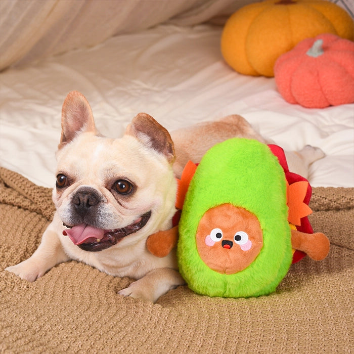 Dog playing with a plush avocado toy on a bed with pumpkins in the background
